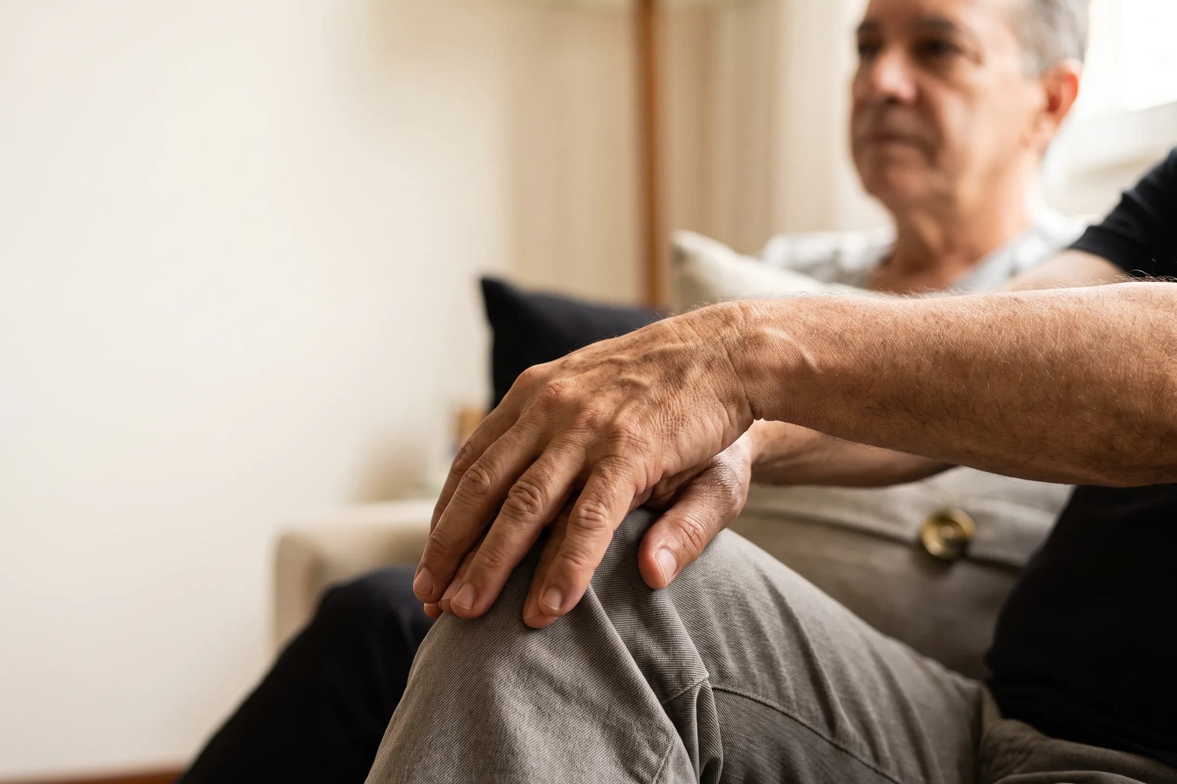 Mãos de uma pessoa mais velha apoiadas sobre o joelho, sentada em ambiente doméstico claro